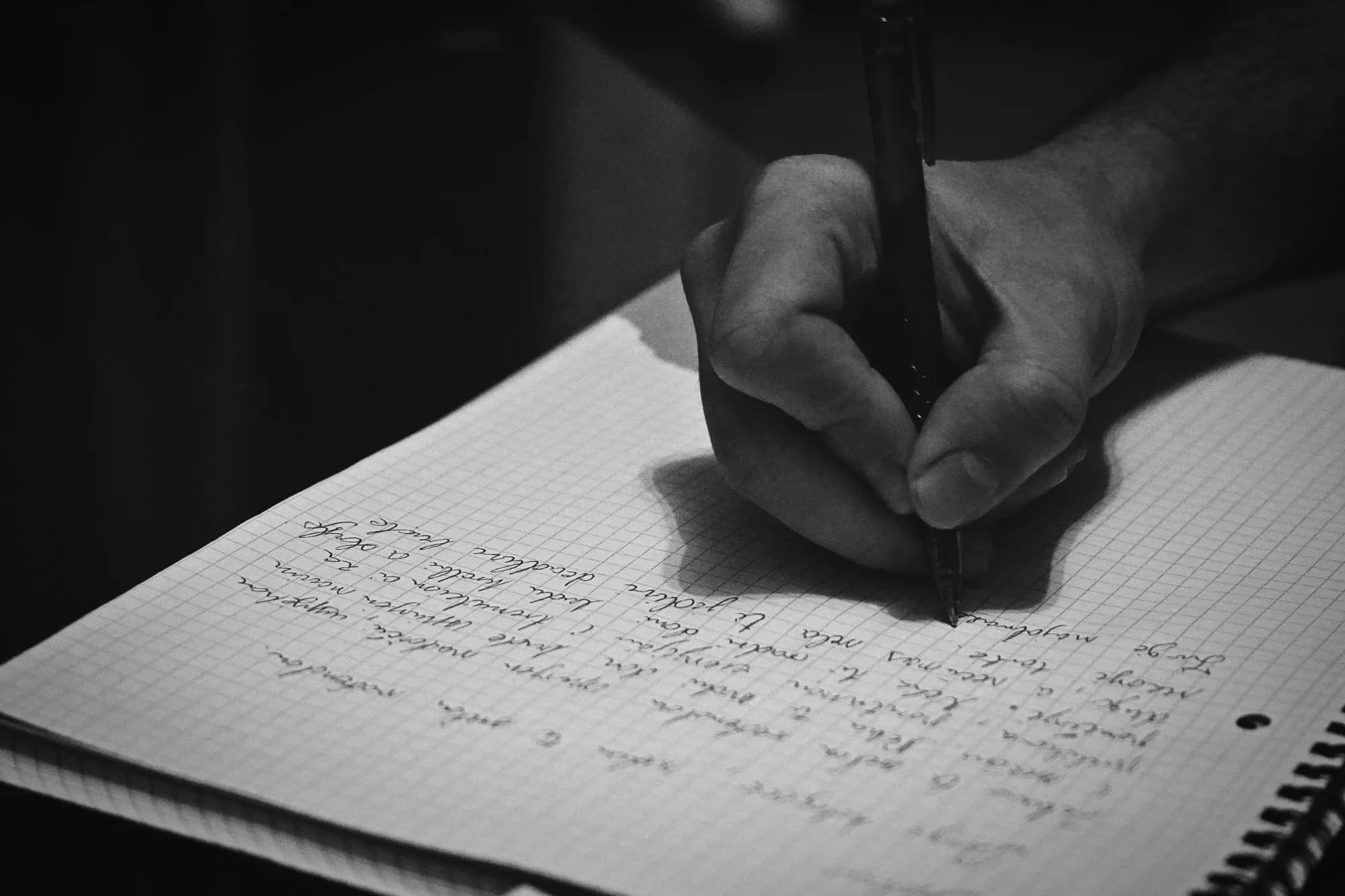 Mano escribiendo con lápiz sobre un cuaderno de hojas cuadriculadas, en blanco y negro.
