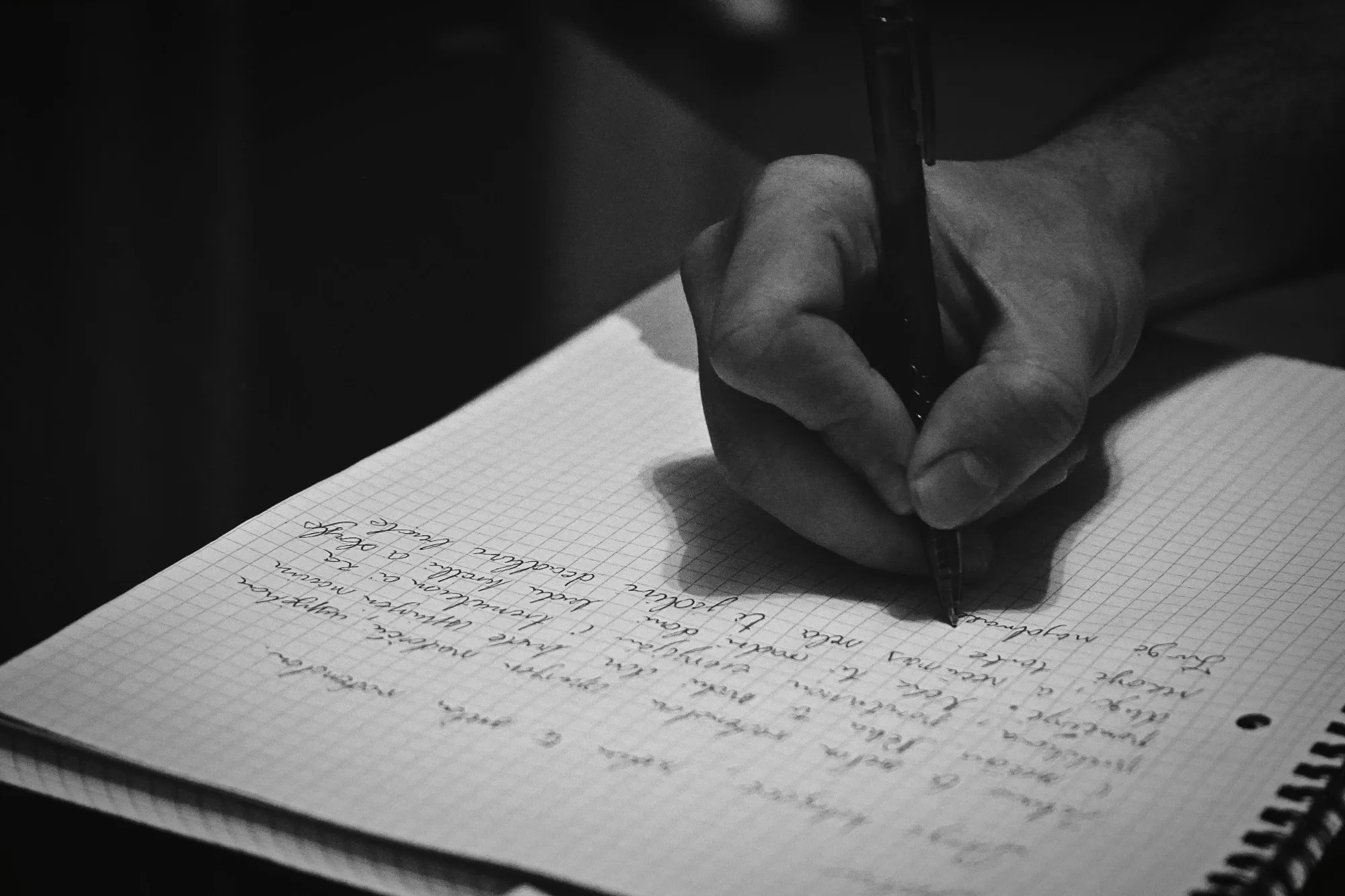 Mano escribiendo con lápiz sobre un cuaderno de hojas cuadriculadas, en blanco y negro.