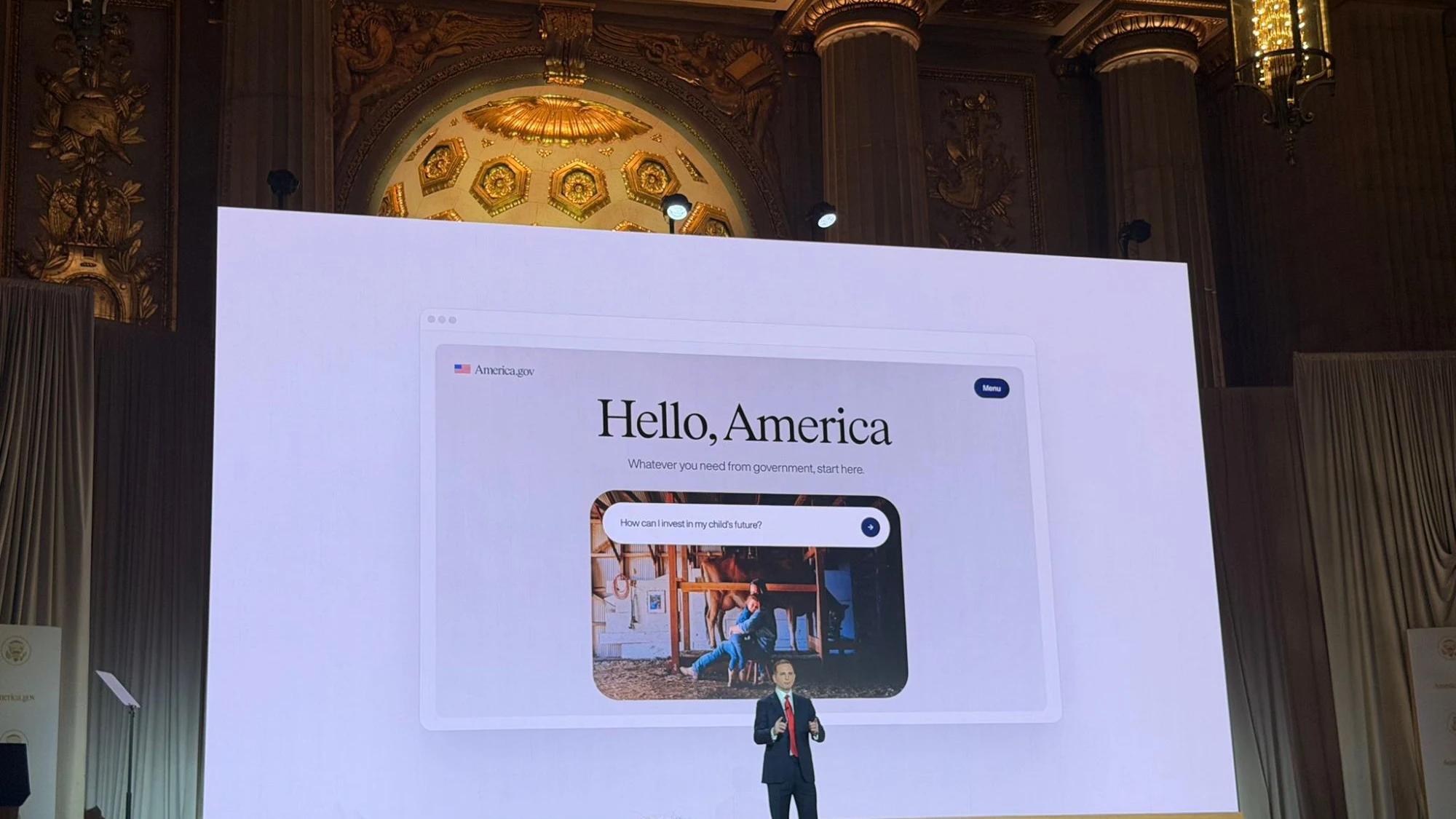 Wide shot inside an ornate auditorium with gilded columns and a chandelier-lit ceiling. A large screen dominates the stage showing the America.gov home page — the words "Hello, America" over a search field asking "How can I invest in my child's future?" A speaker in a dark suit and red tie stands in front of the screen, addressing a seated audience whose silhouettes fill the foreground.